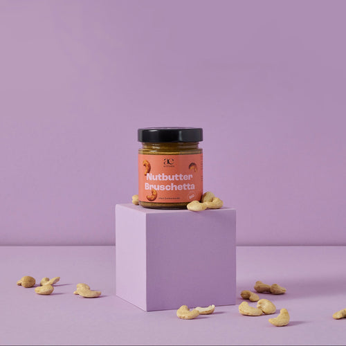 Glas Nutbutter Bruschetta von ae kitchen auf lila Podest, umgeben von Cashews, vor lila Hintergrund