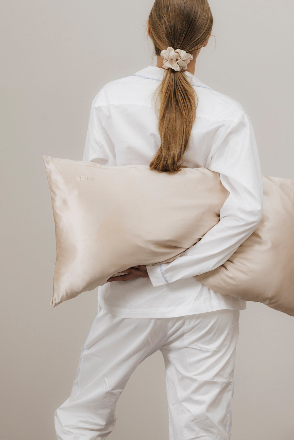 Rückansicht einer Frau in weißem Pyjama, die ein beiges Seidenkissen im Arm hält und einen Zopf mit einem Scrunchie aus Maulbeerseide trägt.