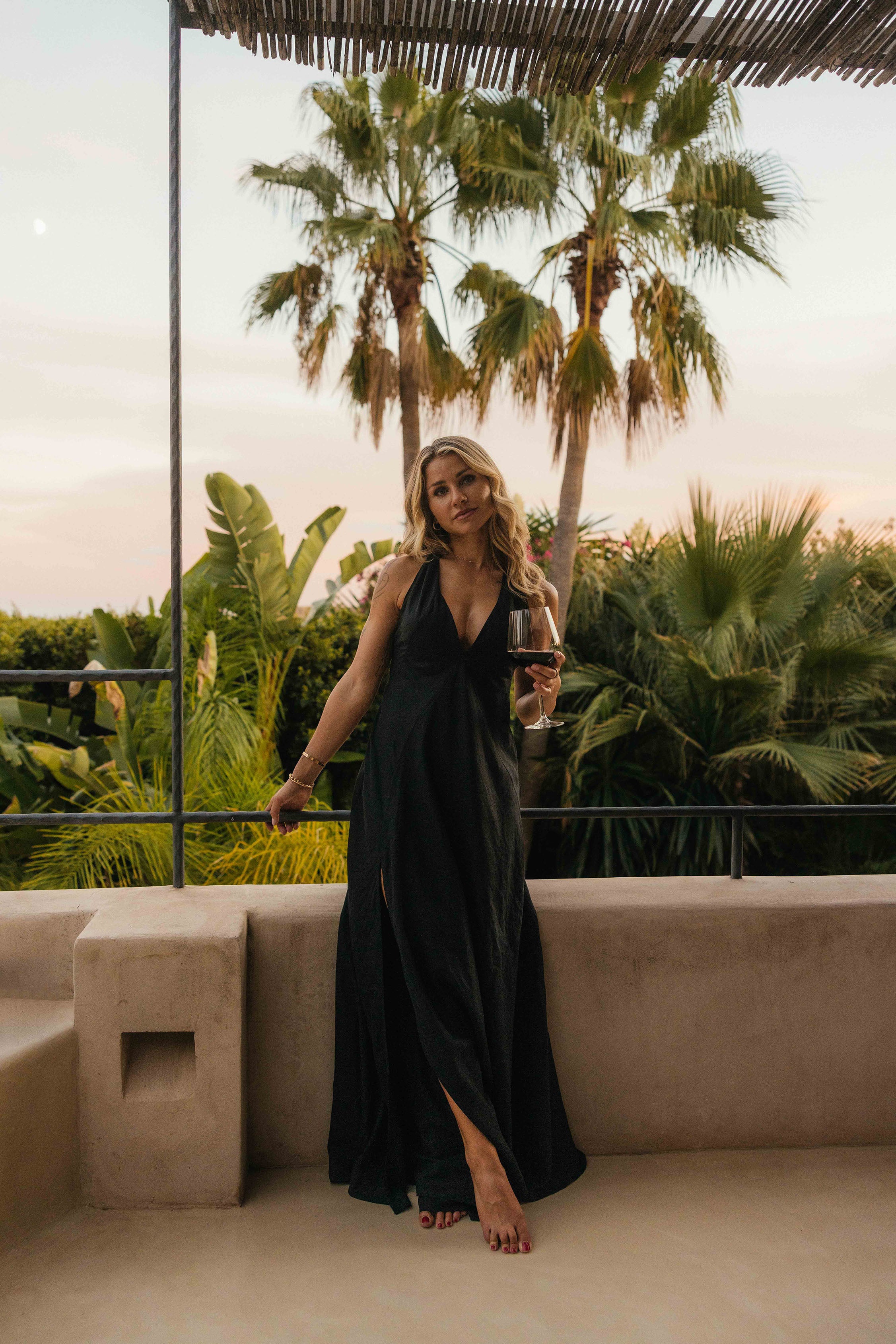 Eine Frau in einem eleganten schwarzen Kleid steht auf einer Terrasse vor tropischen Palmen und hält ein Glas Rotwein in der Hand. Die Abendsonne taucht die Szene in warmes, weiches Licht.