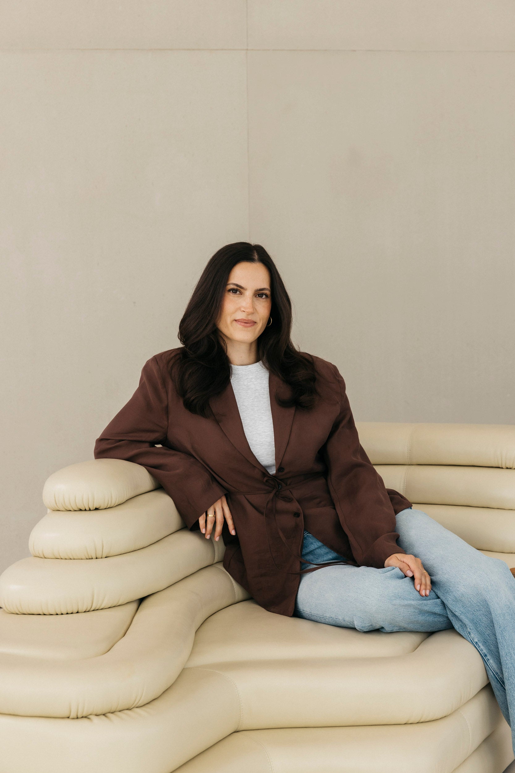 Frau mit dunklen, gelockten Haaren sitzt entspannt auf einem beigen Design-Sofa und trägt einen braunen Blazer mit Jeans.