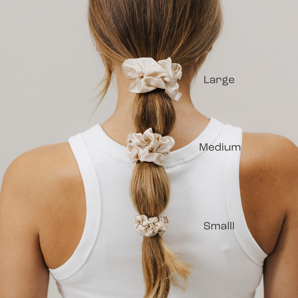 Rückansicht einer Frau mit Pferdeschwanz, der mit drei beigefarbenen Seiden-Scrunchies gestylt ist. Oben ein großes Scrunchie (Large), darunter ein mittleres (Medium) und unten ein kleines (Small). Beschriftung der Größen steht daneben.