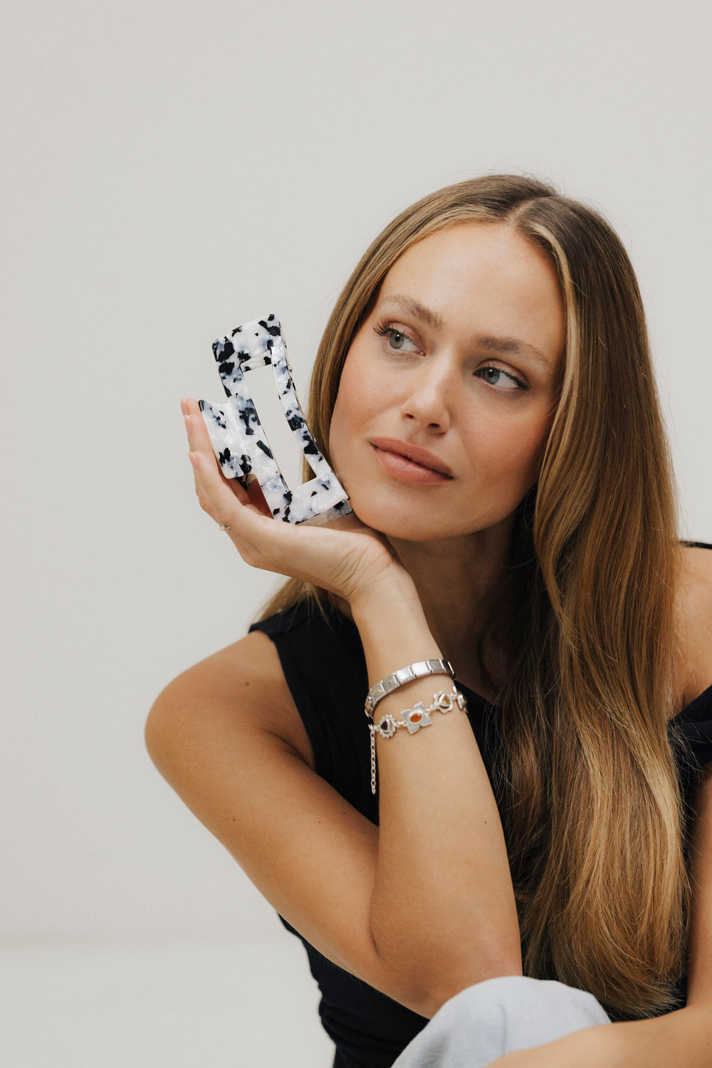 Frau mit langen, glatten, dunkelblonden Haaren hält eine schwarz-weiß gemusterte Haarklammer aus Bio-Acetat an ihr Gesicht und blickt nachdenklich zur Seite.