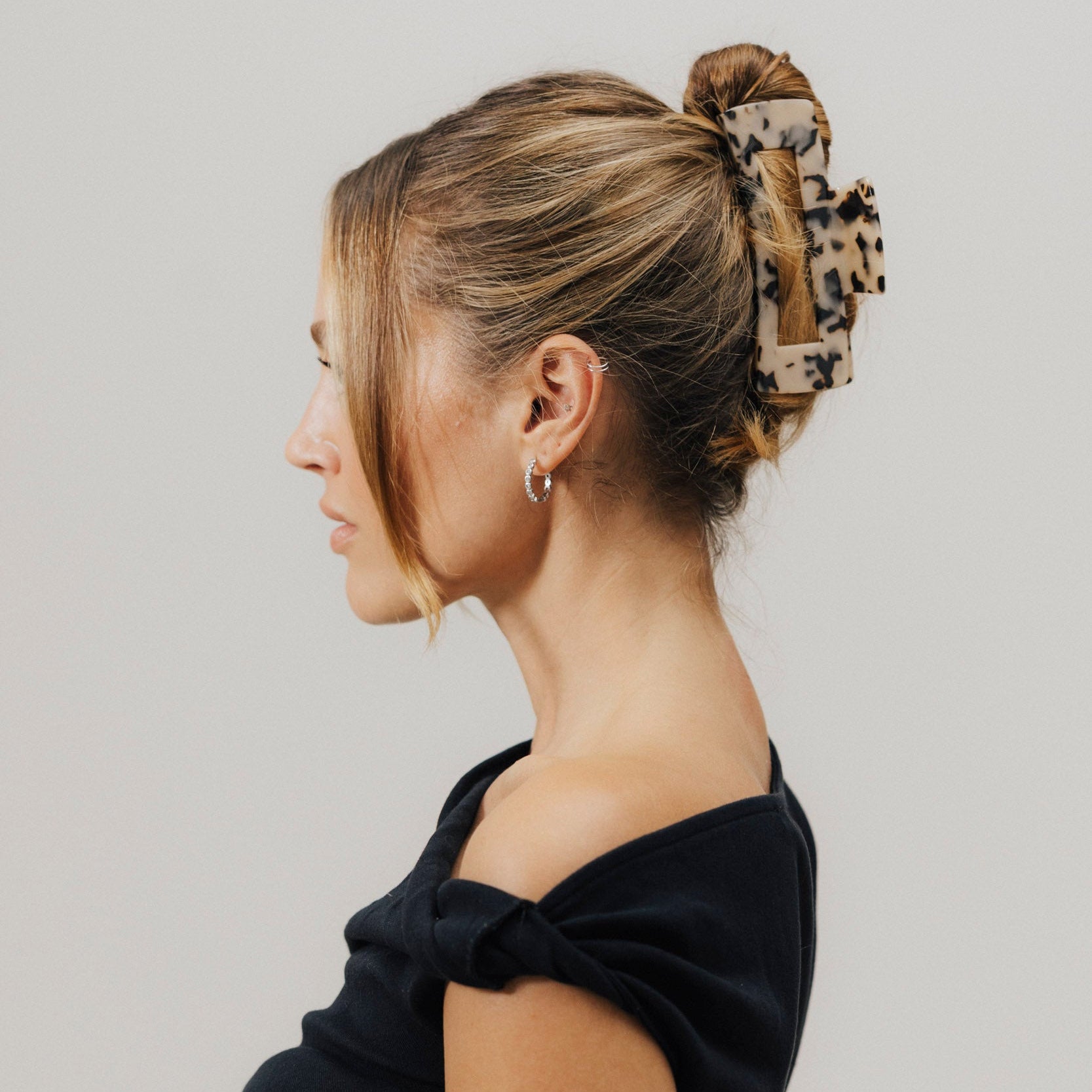 Frau mit hochgestecktem Haar, gehalten von einer großen Haarklammer im Tortoise-Look. Sie trägt ein schwarzes Off-Shoulder-Oberteil und silberne Creolen.