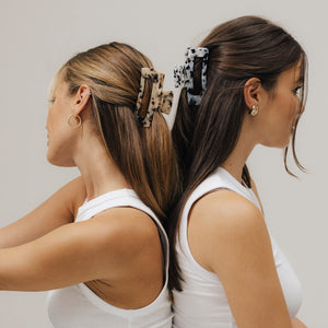 Zwei Frauen sitzen Rücken an Rücken und tragen unsere Haarklammern aus Bio-Acetat – eine in Tortoise, eine in Schwarz-Weiß. Beide tragen weiße Tops und Jeans.