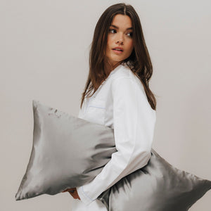 Frau mit braunen Haaren im weißen Pyjama hält ein graues Seidenkissen und blickt über die Schulter zur Seite.