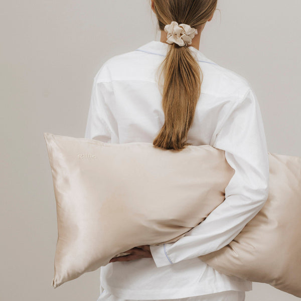 Frau von hinten mit langem Haar im beigen Seiden-Scrunchie, hält einen Kissenbezug aus Maulbeerseide in der Farbe beige – purer Komfort für Nacht & Haar.