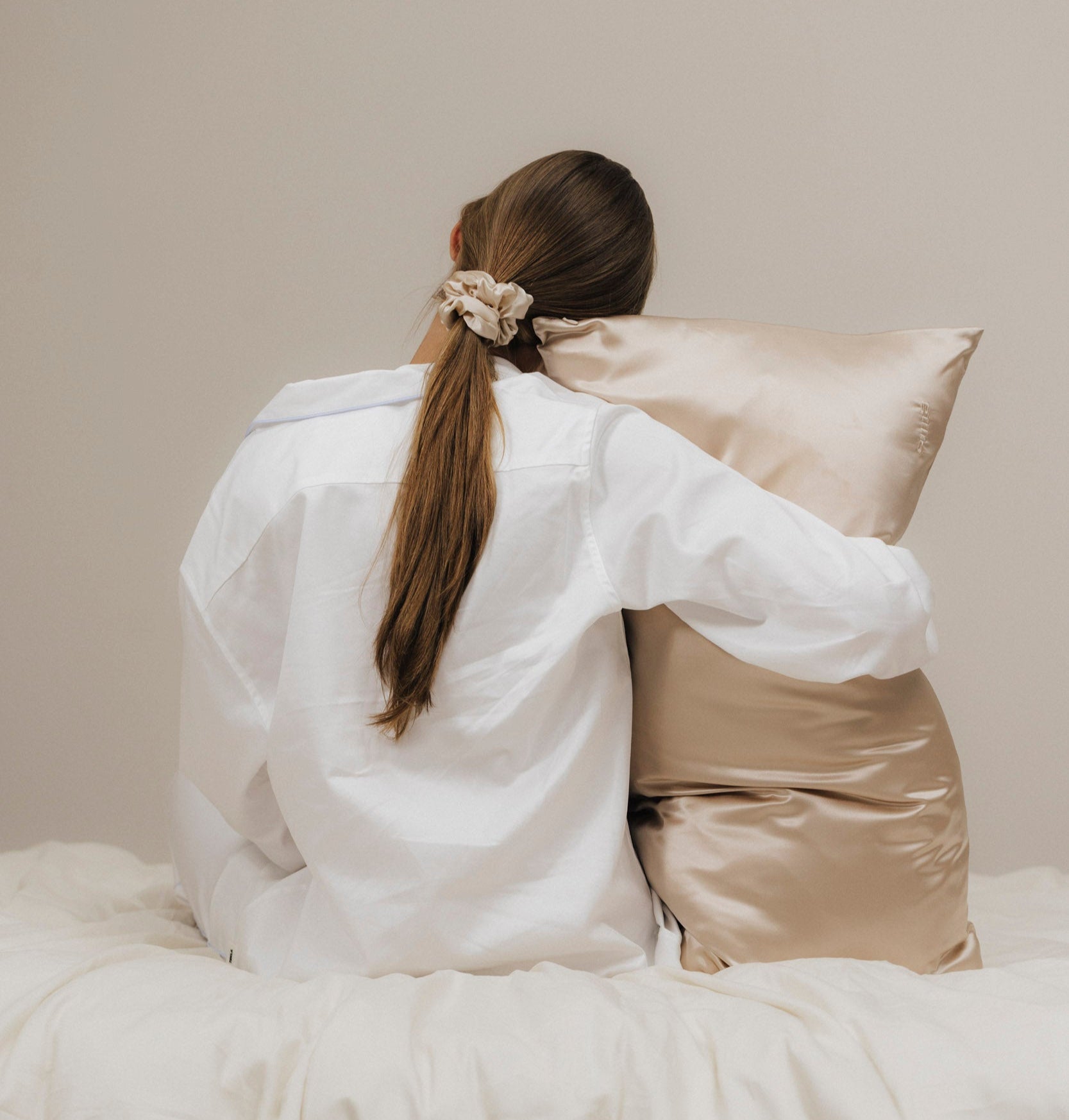 Braunhaarige Frau mit weißen Pyjama, sitzt auf dem Bett und umarmt ein Kissen mit unserem beigen Seidenkissen Bezug.