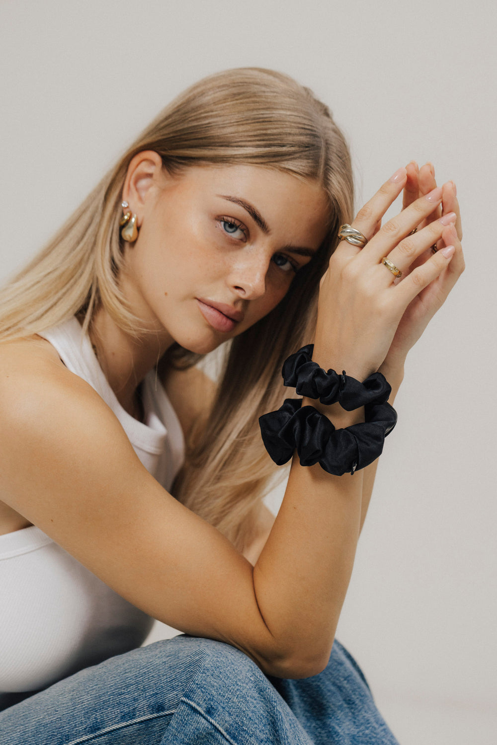 Junge Frau mit langen, blonden Haaren trägt zwei schwarze Scrunchies aus Maulbeerseide ums Handgelenk und schaut ruhig in die Kamera.